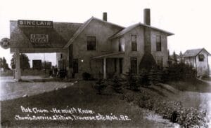 Black-And-White Photo Of A Small Sinclair Gas Station Canopy Beside A House-Like Building, With Pumps Near The Road And Handwritten Text Reading “Ask Chum — He Might Know.”