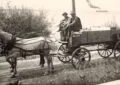 Black-And-White Photo Of A Horse Harnessed To A Wooden Wagon Labeled “Ice Wagon,” With Two Men Seated On The Front Bench On A Dirt Road Near The Shoreline And A Steamer In The Distance.
