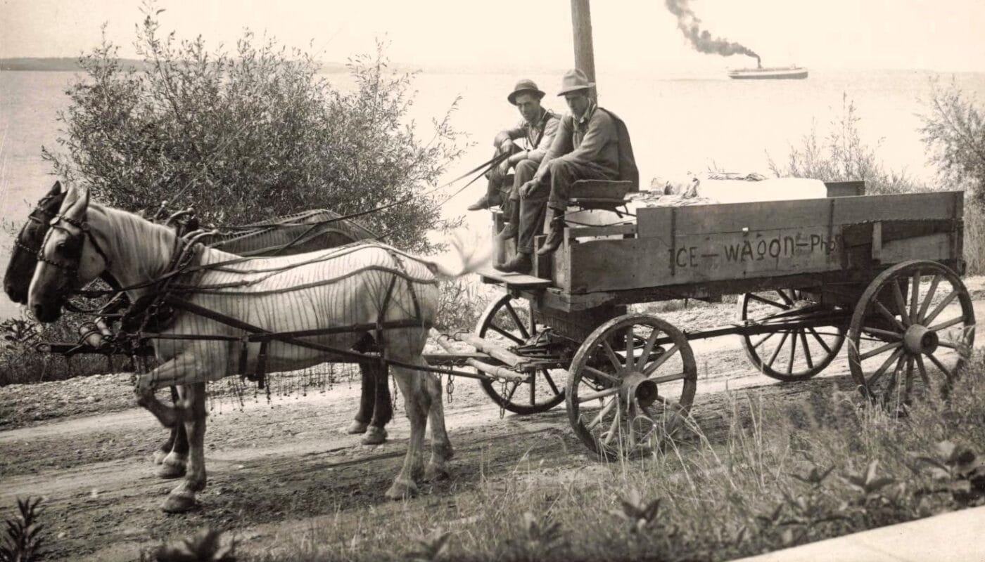 Black-And-White Photo Of A Horse Harnessed To A Wooden Wagon Labeled “Ice Wagon,” With Two Men Seated On The Front Bench On A Dirt Road Near The Shoreline And A Steamer In The Distance.