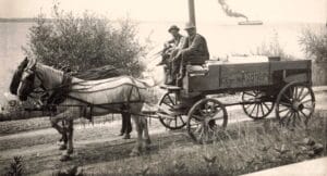 Black-And-White Photo Of A Horse Harnessed To A Wooden Wagon Labeled “Ice Wagon,” With Two Men Seated On The Front Bench On A Dirt Road Near The Shoreline And A Steamer In The Distance.