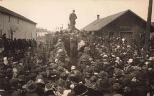 A Dense Crowd Surrounds Uniformed Men Near A Train At A Depot Building Labeled “Shelby,” With People Packed Tightly In The Foreground.