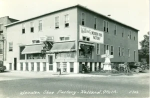 Black-And-White Postcard Photo Of A Two-Story Corner Building Labeled “Holland’s Wooden Shoe Factory,” With Storefront Windows, A Small Windmill Display, Bicycles, And Parked Cars.
