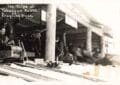 Black-And-White Photo Of People Lined Up Inside A Log Building Beside Several Toboggan Lanes, With A Sled And Riders Blurred As They Speed Past.