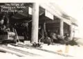 Black-And-White Photo Of People Lined Up Inside A Log Building Beside Several Toboggan Lanes, With A Sled And Riders Blurred As They Speed Past.