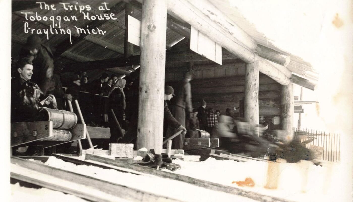 Black-And-White Photo Of People Lined Up Inside A Log Building Beside Several Toboggan Lanes, With A Sled And Riders Blurred As They Speed Past.