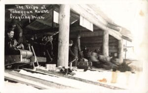 Black-And-White Photo Of People Lined Up Inside A Log Building Beside Several Toboggan Lanes, With A Sled And Riders Blurred As They Speed Past.