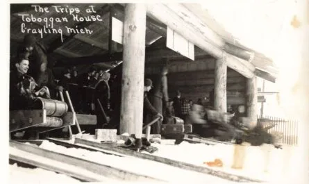Black-And-White Photo Of People Lined Up Inside A Log Building Beside Several Toboggan Lanes, With A Sled And Riders Blurred As They Speed Past.