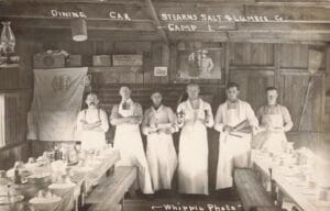 Six Men In Aprons Stand In A Wooden Dining Hall Labeled “Dining Car Stearns Salt &Amp; Lumber Co. Camp 1,” With Long Tables Set For A Meal On Both Sides.