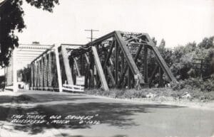 Black-And-White Photo Of Several Adjacent Steel Truss Bridges Over A River In Blissfield, Michigan, Viewed From A Road Approach With Trees In The Background.