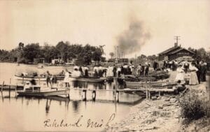 A Large Crowd Gathers Beside A Lakeside Depot Labeled “Lakeland” While Several Small Boats Wait At Wooden Docks In The Foreground.