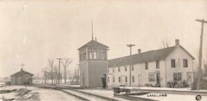 Black-And-White Winter Photo Of The Lakeland Depot Area With Curving Railroad Tracks, A Tall Tower With A Man In The Doorway, And A Nearby Building Labeled “Post Office.”