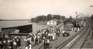 Black-And-White Image Of A Crowded Lakeside Depot Platform At Lakeland, Michigan, With A Building Labeled “Lakeland” Beside Railroad Tracks And Water.