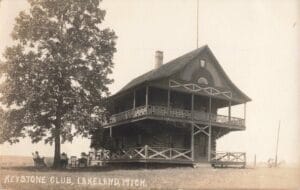 A Black-And-White Photo Of A Log Clubhouse With A Wraparound Porch And Decorative Railings, With Several People Sitting In Chairs Under A Large Tree At Left.