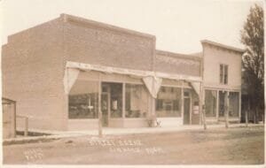 A Sepia Street View Of Two Connected Storefront Buildings With Large Display Windows, Fabric Awnings, And “Street Scene Linwood Mich.” Handwritten At The Bottom.