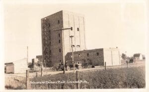 A Tall Brick Industrial Building Stands In A Rural Field With Two Vintage Cars Parked In Front, And Handwritten Text At The Bottom Reads “Schanzer Chicory Plant, Linwood, Mich.”