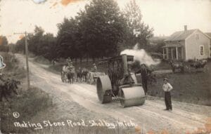 Black-And-White Photo Labeled “Making Stone Road, Shelby, Mich.” Showing A Steam Road Roller On A Rural Road With Workers, Horse-Drawn Wagons, And Houses In The Background.