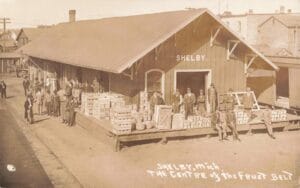 Sepia-Toned Image Of The Shelby Railroad Depot With Men Standing On A Platform Piled With Fruit Baskets And Crates Beside Train Tracks.