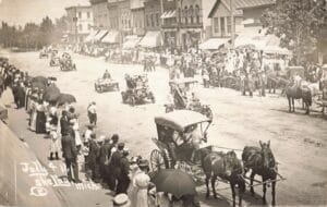 A Crowded 1911 Downtown Parade Scene In Shelby, Michigan, With Early Automobiles And A Horse-Drawn Team On A Wide Dirt Street And Spectators Lining Both Sidewalks.
