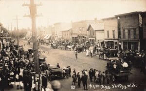 A Sepia Street Scene In Shelby, Michigan, Crowded With People, Early Automobiles, A Few Horse-Drawn Vehicles, And Storefronts, With “$1.00 Day, Shelby, Mich.” Written At The Bottom.