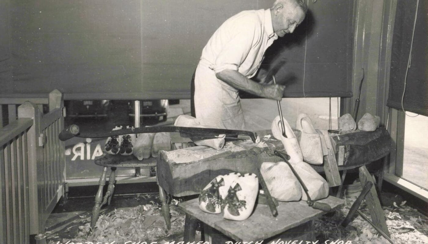 Black-And-White Postcard-Style Image Of A Corner Building Labeled “Holland’s Wooden Shoe Factory,” With Large Shop Windows, People Gathered Near The Entrance, And Cars Parked Along The Street.