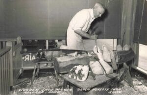 Black-And-White Postcard-Style Image Of A Corner Building Labeled “Holland’s Wooden Shoe Factory,” With Large Shop Windows, People Gathered Near The Entrance, And Cars Parked Along The Street.