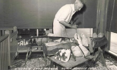Black-And-White Postcard-Style Image Of A Corner Building Labeled “Holland’s Wooden Shoe Factory,” With Large Shop Windows, People Gathered Near The Entrance, And Cars Parked Along The Street.