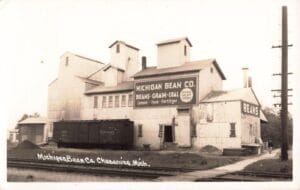 A Black-And-White Photo Of The Michigan Bean Co. Elevator Complex In Chesaning With A Large Wall Sign, A Railcar Beside The Building, And Railroad Tracks In The Foreground.