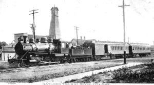 Black-And-White Postcard Image Of A Steam Locomotive With A Tender Marked “11” Pulling Passenger Cars Beside A Depot Area In Boyne City, Michigan.