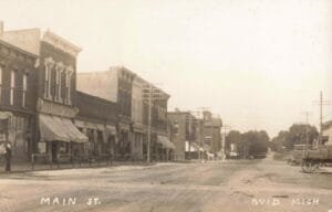 A Sepia-Toned Street Scene Labeled “Main St. Ovid Mich,” Showing Brick Storefronts With Awnings On The Left, A Wide Dirt Road, Utility Poles, And A Horse-Drawn Wagon At Right.