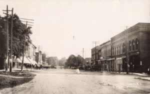 Wide View Down A Dirt Main Street Lined With Two-Story Storefronts And Awnings, With Utility Poles, Horse-Drawn Wagons And Early Automobiles Parked Along Both Sides.