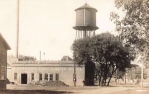 A Brick Building Labeled “Ovid Electric Light &Amp; Water Works” Sits Beside Railroad Tracks With A Tall Wooden Water Tank On A Steel Tower Behind Trees.