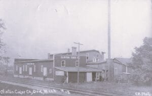 A Large Industrial Building Labeled “Clinton Cider Co.” Stands Beside Railroad Tracks In Ovid, Michigan, With A Tall Smokestack And Multiple Connected Sections.