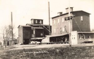 Black-And-White Photo Of The Ovid Roller Mills Complex With Tall Mill Buildings, Two Smokestacks, A Utility Pole With Wires, And A Horse-Drawn Wagon Near A Loading Dock.