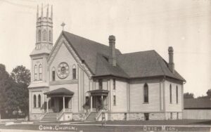 A Large Wooden Church With A Tall Spired Tower, Arched Windows, And A Circular Rose Window Stands In Ovid, Michigan, With Steps And Small Porches At The Entrances.