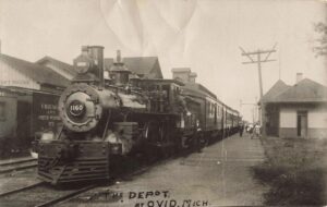 Black-And-White Photo Of Steam Locomotive No. 1160 Stopped Beside A Depot Platform In Ovid, Michigan, With Several People On The Platform And Passenger Cars Trailing Behind.