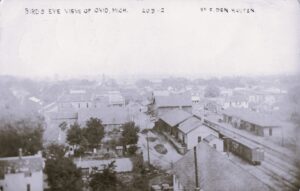 A Hazy Elevated View Over Ovid, Michigan, Showing Clustered Buildings, Dirt Streets, Trees, And Railroad Tracks With A Freight Car On The Right.