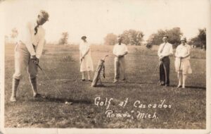 A Vintage Photo Shows Four Golfers On A Grassy Course, With One Man Preparing To Hit And Three Others Watching Near A Standing Golf Bag.