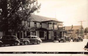 Black-And-White Street Scene Showing The Two-Story Mowry Hotel And Restaurant With Porch Columns And Several Late-1930S Cars Parked In Front, With A “Groceries And Meats” Storefront Beside It.