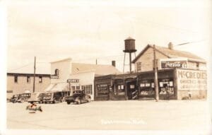 A Vintage Photo Of Downtown Roscommon, Michigan, Shows Several Storefronts With Signs For Mccredie’s Groceries &Amp; Meats, Beanie’s, And A Soda/Drugs Shop, With Parked Cars And A Water Tower Behind The Buildings.