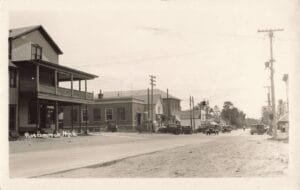 Black-And-White Photo Looking Down A Wide Street In Roscommon, Michigan, With Several Early Cars Parked Along The Curb And A Large Two-Story Building With Wraparound Porches On The Left.