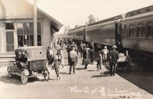 Black-And-White Photo Of A Crowded Depot Platform Beside Passenger Train Cars, With People, Luggage, A Handcart And An Early Automobile Near The Station Building.