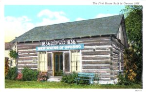 A Color Postcard Shows A Small Log Cabin Labeled As Owosso’s First House Built In 1836, With Signs Over The Doorway Reading “Comstock’s Home” And “Beginning Of Owosso.”