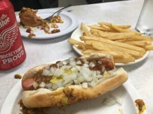 A Coney Dog Topped With Chili, Yellow Mustard And Chopped Onions Sits On A Plate Beside French Fries And A Can Of Labatt Blue.