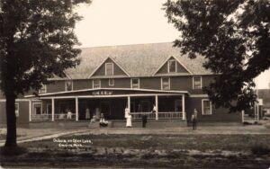 Sepia-Toned Photo Of A Large Two-Story Wooden Inn Labeled “Ockers,” With People Standing And Sitting On The Front Porch, Framed By Tre