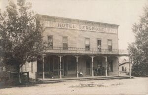 Black-And-White Photo Of A Two-Story Wooden Building Labeled “Hotel Densmore,” With Several People Sitting And Standing On The Porch And Balcony Beside A Dirt Road.