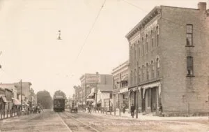 A Sepia Street Scene Shows An Interurban Railcar On Tracks Down Main Street With People, Wagons, And A Tall Brick Building Marked “Union Block” On The Right.