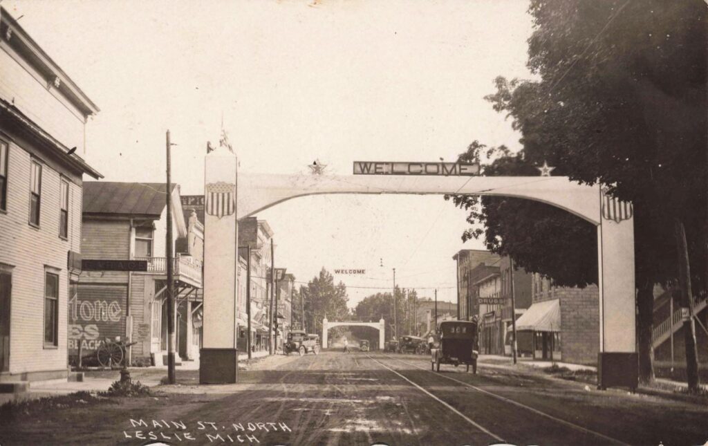 Early 1900S Main Street Leslie Michigan With Welcome Arch, Storefronts, And Dirt Road Traffic.