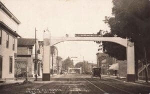 Early 1900S Main Street Leslie Michigan With Welcome Arch, Storefronts, And Dirt Road Traffic.