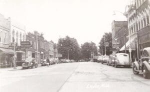 Black-And-White View Down A Wide Main Street In Leslie, Michigan, With Parked Vintage Cars On Both Sides And Storefront Signs Including “Morea’s Restaurant” And Appliance Advertising.