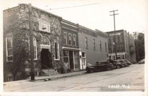 A Stone-Front Building Labeled “G.a.r. Hall” Stands Along A Street In Leslie, Michigan, With Several Vintage Cars Parked In Front And More Buildings Stretching Down The Block.